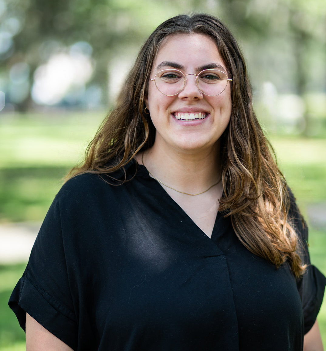 Smiling woman with glasses and long brown hair wearing black scrubs outdoors