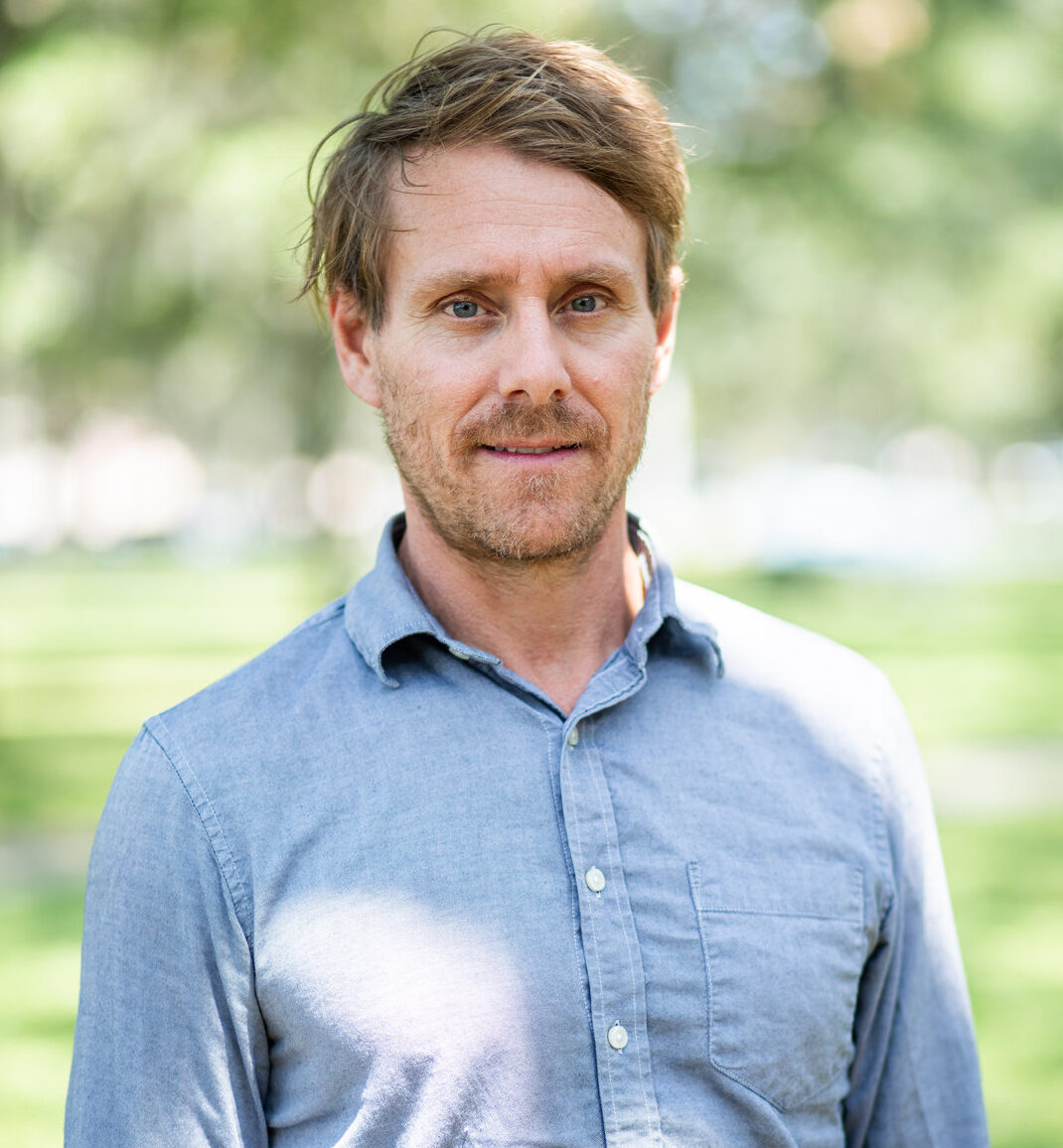 Professional headshot of smiling man in blue button-down shirt outdoors