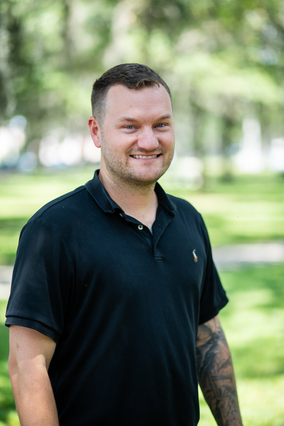 Smiling man in black polo shirt with tattoo standing outdoors in green park setting