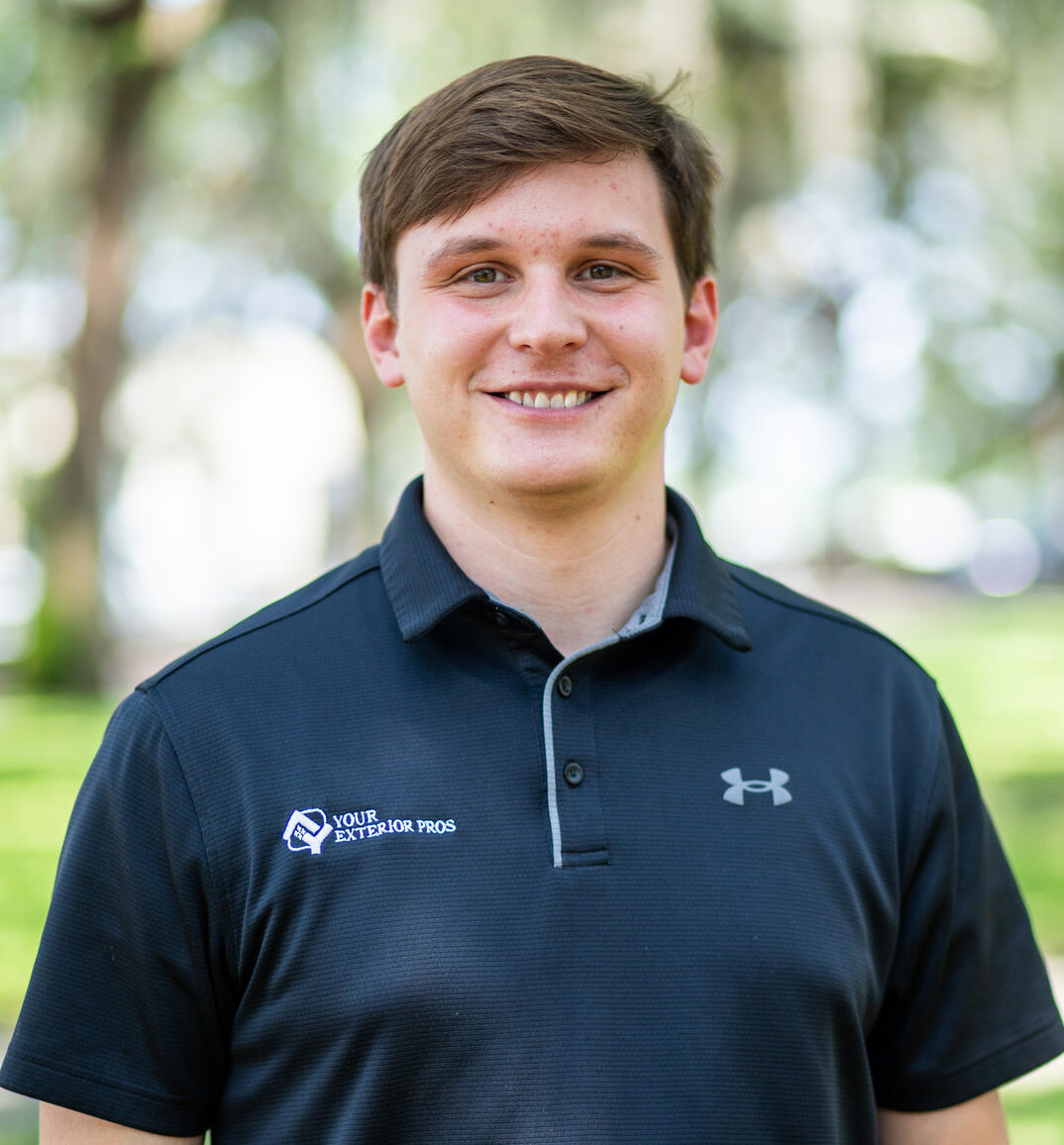 Young man smiling in navy Under Armour polo shirt outdoors with green trees